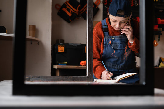 Female worker working as handyman or mechanic in furniture repair workshop making phone call using smartphone