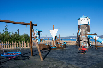 Fototapeta premium A nautical-themed children’s playground with a play ship, lighthouse slide, and swing sits near a scenic waterfront.