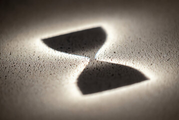 Dramatic close-up of an hourglass shape formed by strong light and shadow on a rough, granular textured surface, symbolizing time's passage.