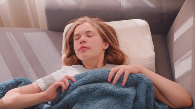 Close up of elegant lady waking up finally stretching herself on couch, resting with soft blanket and pillow as morning sunlight streams through curtains creating cozy peaceful atmosphere