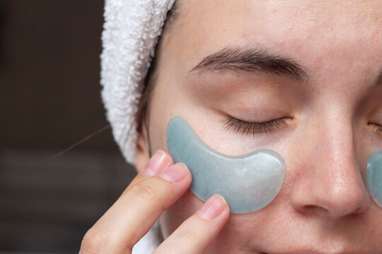 Horizontal photo close up. Woman with closed eyes, white towel on head applies blue eye patches to face in hotel bed room. Concept of beauty, make up, relax, health, spa, rest, skin care