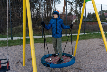 Boy in blue jacket standing and balancing on nest swing holding ropes at playground on autumn day. Concept of childhood balance skills, adventurous outdoor play and active physical recreation for kids