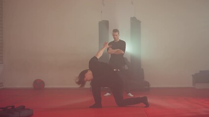 Karate practitioner performing dynamic movement during sparring training in gym, arms raised in motion on red mats, opponent standing in background, martial arts discipline with focus, under bright