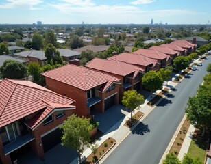 Obraz premium Aerial view shows modern brick townhouses lining suburban street. Newly built homes with red roofs create neat row. Distant city buildings dot horizon in Melbourne Australia residential area during