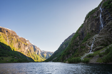Scenic view of a majestic Norwegian fjord with steep green mountains, cascading waterfalls, and calm water on a sunny summer day.