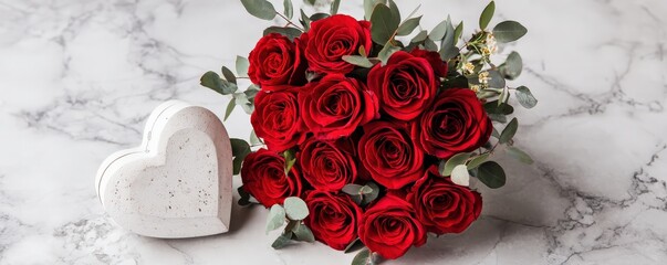 A bouquet of red roses arranged in a heart shape beside a decorative heart, set against a marble background.