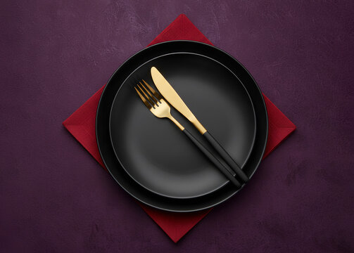 A matte black plate and gold cutlery resting on a red napkin with minimalist table setting against a dark textured surface. Christmas and New Year festive ambiance for holiday dining. - Powered by Adobe