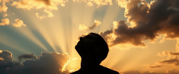 Silhouette of a man looking up at a dramatic sky with sun rays. Hope, faith, and spirituality concept. Backlit person against a golden sunset background