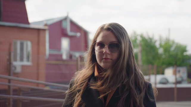 serene woman in sunglasses turns toward camera wearing brown scarf under dark coat with loose hair lifted by breeze, morning light over suburban houses and modern street quiet residential vibe