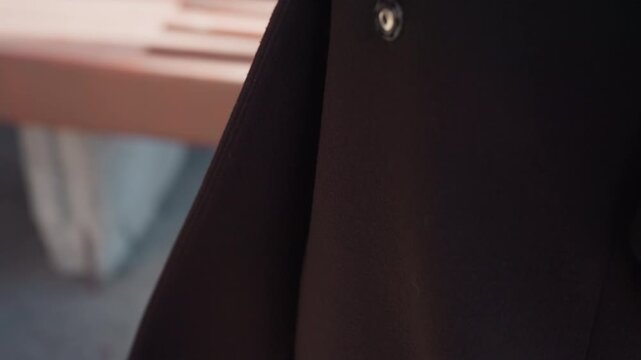 tight detail shows wool long coat swaying at hem, shiny snap buttons catching sunlight, soft bench in background, street mood, minimalist fashion vibe, muted colors and gentle breeze moving fabric
