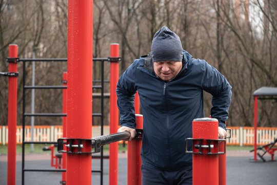 Focused man exercise on parallel bars in an urban outdoor gym after rain. Dressed warmly, concentrating on fitness routine amidst city backdrop, emphasizing strength and determination. Daily routine - Powered by Adobe