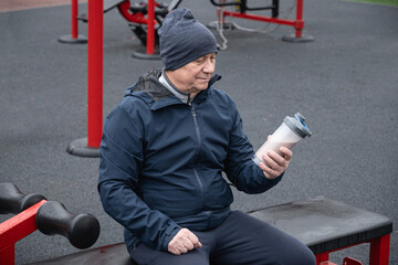 Adult man sits on an outdoor gym bench holding protein shake, wearing casual vest. He relaxed,...