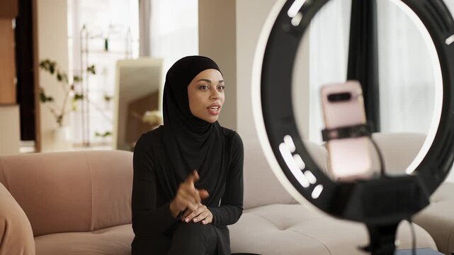 Smiling Arab woman in hijab recording lifestyle vlog on smartphone at home