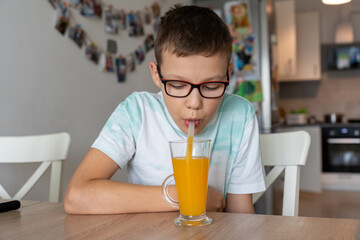 Boy with glasses drinking fresh homemade orange juice through straw at kitchen table at home....