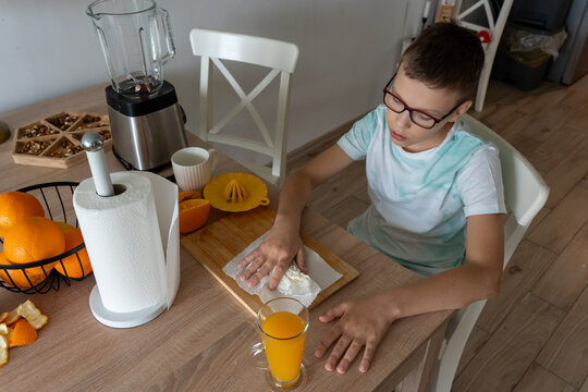 Boy with glasses wiping hands with paper towel after making fresh orange juice at kitchen table at home. Concept of children hygiene habits, cleaning up after cooking and teaching tidy routine.