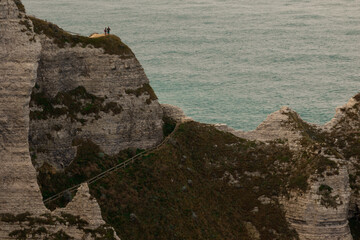 A couple stands on a high rocky cliff overlooking the calm blue sea, surrounded by rugged coastal formations and soft evening light.