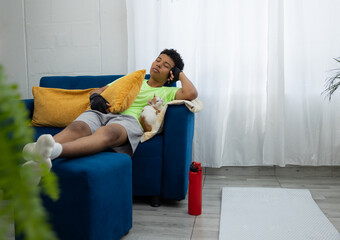 Brown skinned teenager asleep on the couch after his home workout, while his cat rests beside him. A warm scene that reflects relaxation after exercising at home