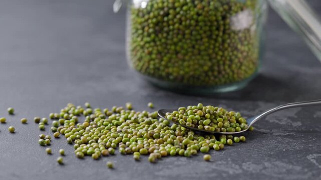 green mung beans in a metal spoon on a dark kitchen table. green mung beans in the glass jar and spilling out to on a dark kitchen table.