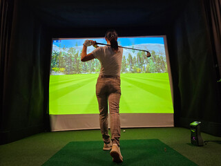 Woman practicing golf swing indoors with virtual screen in the background at a simulation center...