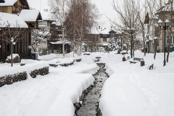 A picturesque pedestrian walkway in the mountain town of Bansko, Bulgaria covered in fresh snow, with a central creek, traditional houses, and bare trees on a cold winter day.