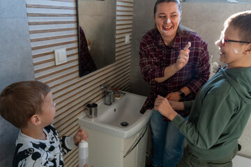 Happy mother and two sons with cream on faces laughing together in bathroom during morning skincare routine near sink. Concept of family fun, parenting joy and teaching children healthy hygiene habits