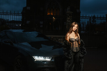 A photograph, a portrait of a beautiful young sexy brunette woman, a stylish model in lingerie, in a fur coat, standing at night in the dark with a chic expensive black car on a city street.