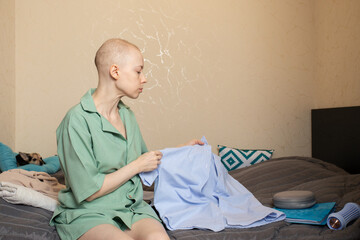 Woman with short hair after cancer treatment sitting on bed at home, preparing clothes for a...