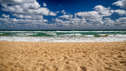 Waves rolling onto a sandy beach under a sky full of clouds on a bright day with turquoise sea and soft foam
