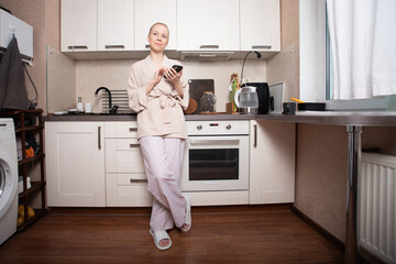 Young woman with cancer using smartphone in cozy home kitchen, bald female patient in bathrobe leaning on countertop, texting with family and 