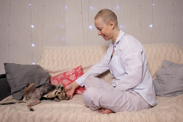smiling oncology patient in loose pyjamas sits cross legged on a cozy sofa, calmly reaching out to stroke her little dog, concept of home comfort, mental health support,