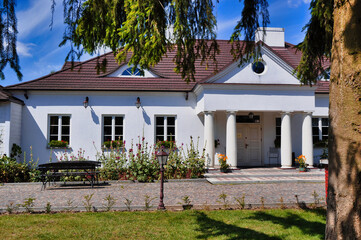 Urokliwy biały zabytkowy dworek z klasycznymi kolumnami, czerwonym dachem, brukowaną ścieżką i przytulnym ogrodem - Soplicowo Cichowo. © Pol Badger