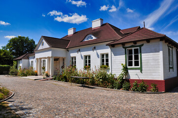 Urokliwy biały zabytkowy dworek z klasycznymi kolumnami, czerwonym dachem, brukowaną ścieżką i przytulnym ogrodem - Soplicowo Cichowo. © Pol Badger