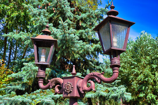 Zabytkowa, ozdobna lampa uliczna z dwiema latarniami i dekoracyjnymi elementami metalowymi, na tle bujnej zieleni i jasnobłękitnego nieba.