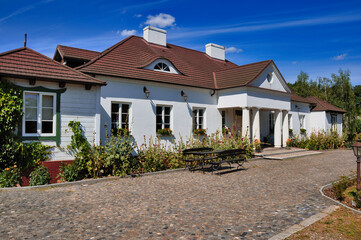 Urokliwy biały zabytkowy dworek z klasycznymi kolumnami, czerwonym dachem, brukowaną ścieżką i przytulnym ogrodem - Soplicowo Cichowo. © Pol Badger