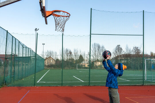 Boy in blue jacket and pompom hat throwing basketball at hoop on outdoor court with soccer field and fence on clear autumn day. Children sports practice, shooting skills and active lifestyle.