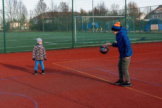 Older brother dribbling basketball while little boy watches on outdoor sports court with soccer goal and playground on autumn day. Concept of siblings playing together, children sports and active fun.