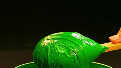 A wooden spatula lifts thick, glossy green ink from a white bucket, emphasizing the rich pigment and sticky texture that ensures even application during manual fabric screen printing. - Powered by Adobe