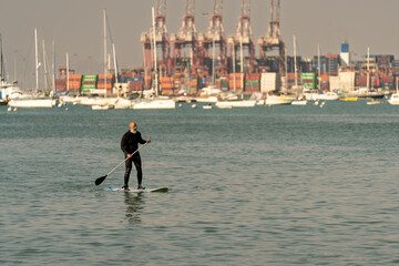 Adult man paddle boarding in bay with industrial port
