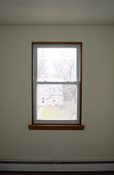 View looking out a single hung window with wooden trim from inside an empty room with white walls