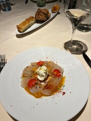 Gourmet White Fish Carpaccio or Ceviche Served on a Minimalist Plate with Citrus Dressing, Edible Flowers, Spices, and a Glass of White Wine in Fine Dining Setting