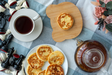 Cottage cheese pancakes on plate and cutting board with tea cup, glass teapot, chess pieces and flowers on checkered tablecloth top view. Concept of cozy breakfast, leisure time and homemade food.