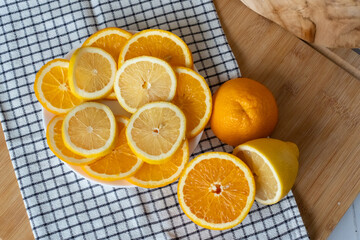 Sliced oranges and lemons on white plate with whole citrus fruits on checkered napkin and wooden cutting board top view. Concept of healthy food preparation, vitamin fruits and kitchen cooking scene.
