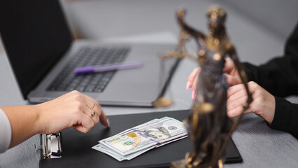 Two hands negotiating over cash on a black folder, with a laptop and a statue of justice in the...