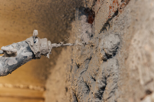 Close-up of a construction worker applying wet cement or mortar (stucco/plaster) to a brick wall using a spray nozzle, creating a rough, uneven texture.
