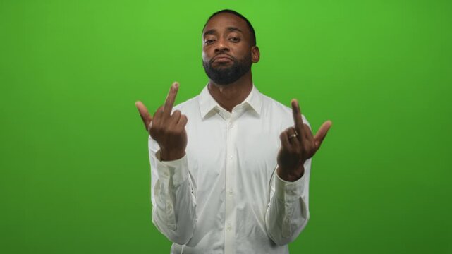 Man showing middle fingers to camera in green studio wearing a white shirt with visible beard and direct gaze; defiance.