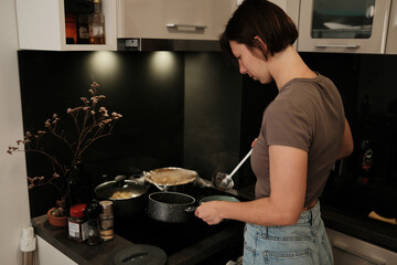 A woman prepares food in a modern kitchen, carefully ladling hot soup into a bowl as part of her daily cooking routine.