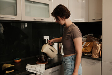 A woman pours hot water from an electric kettle into a glass teapot, preparing a warm drink in a calm kitchen setting.