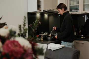 A woman cooks in a cozy kitchen, stirring food in a pot under soft lights and creating a warm, calm everyday atmosphere.