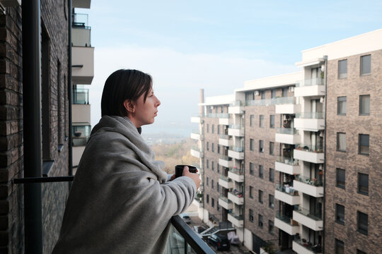 A woman leans on a balcony railing with a warm drink, quietly observing the cityscape in soft daylight.