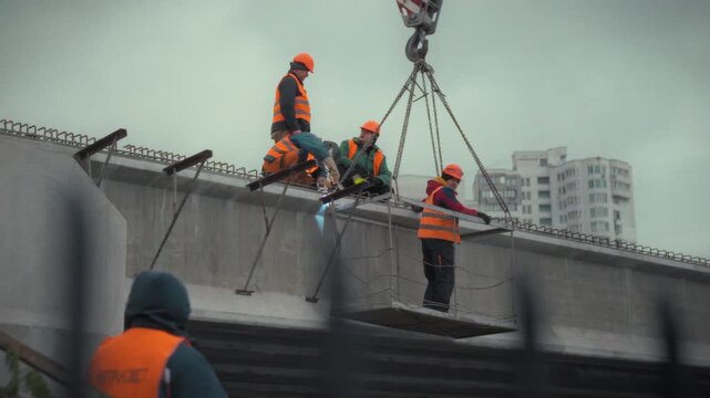 Construction workers wearing hard hats and safety vests perform repairs on a concrete road bridge. They are working together at a height, ensuring safety and precision in their tasks